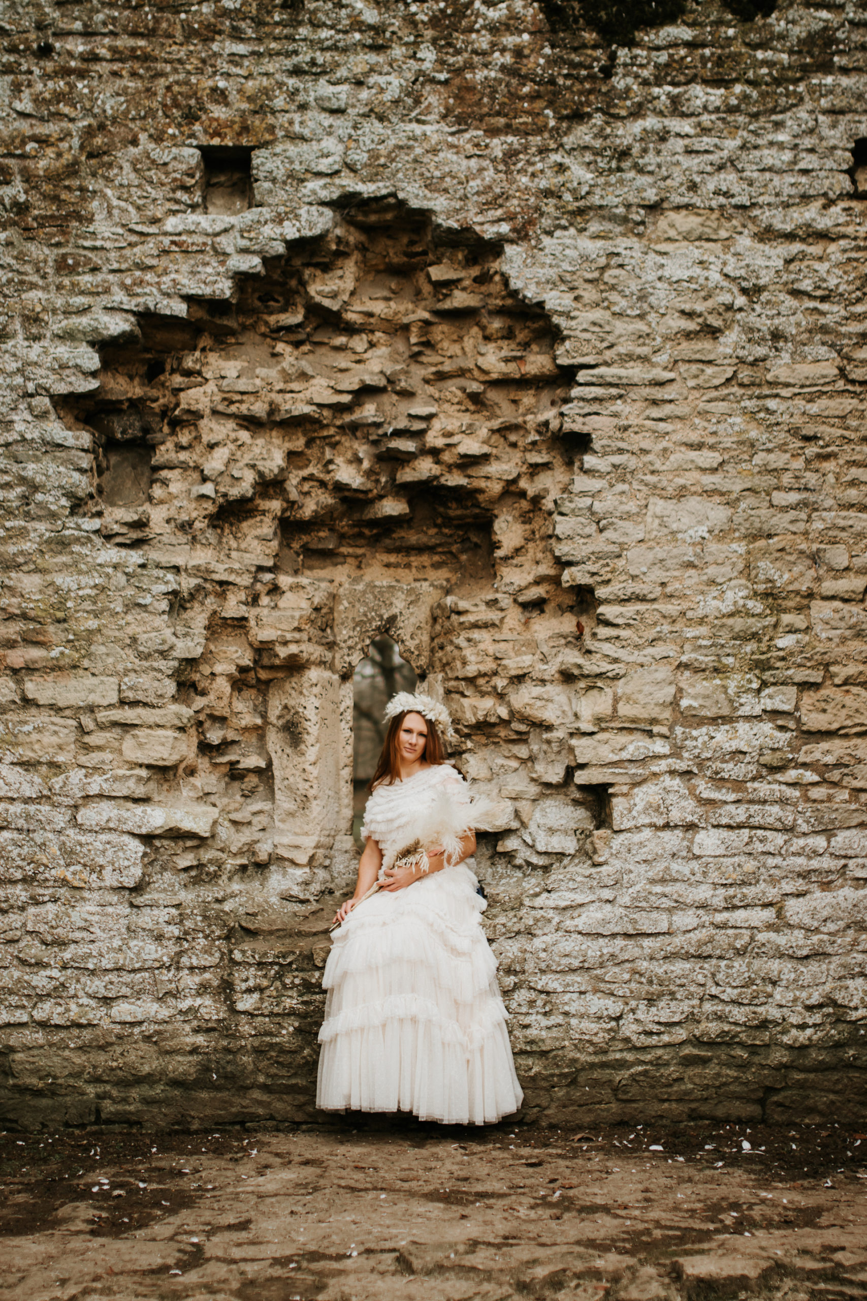 Bride wearing ruffled needle and thread wedding dress, sat down on ledge in Minster Lovell Ruins | Bridal Portraits in The Cotswolds