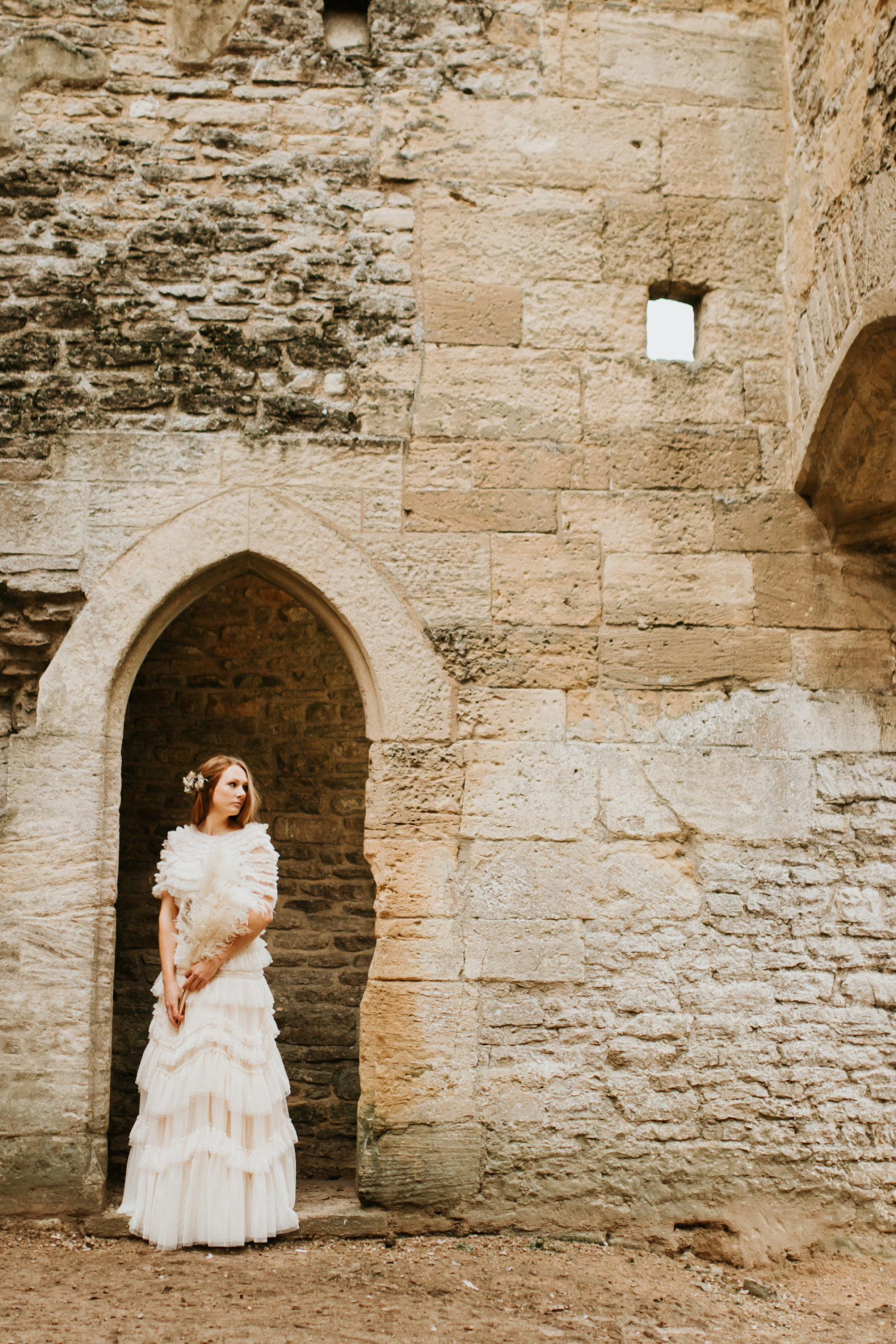 Bride wearing ivory ruffled bridal gown, holding dried flower bouquet and standing in Minster Lovell Ruins