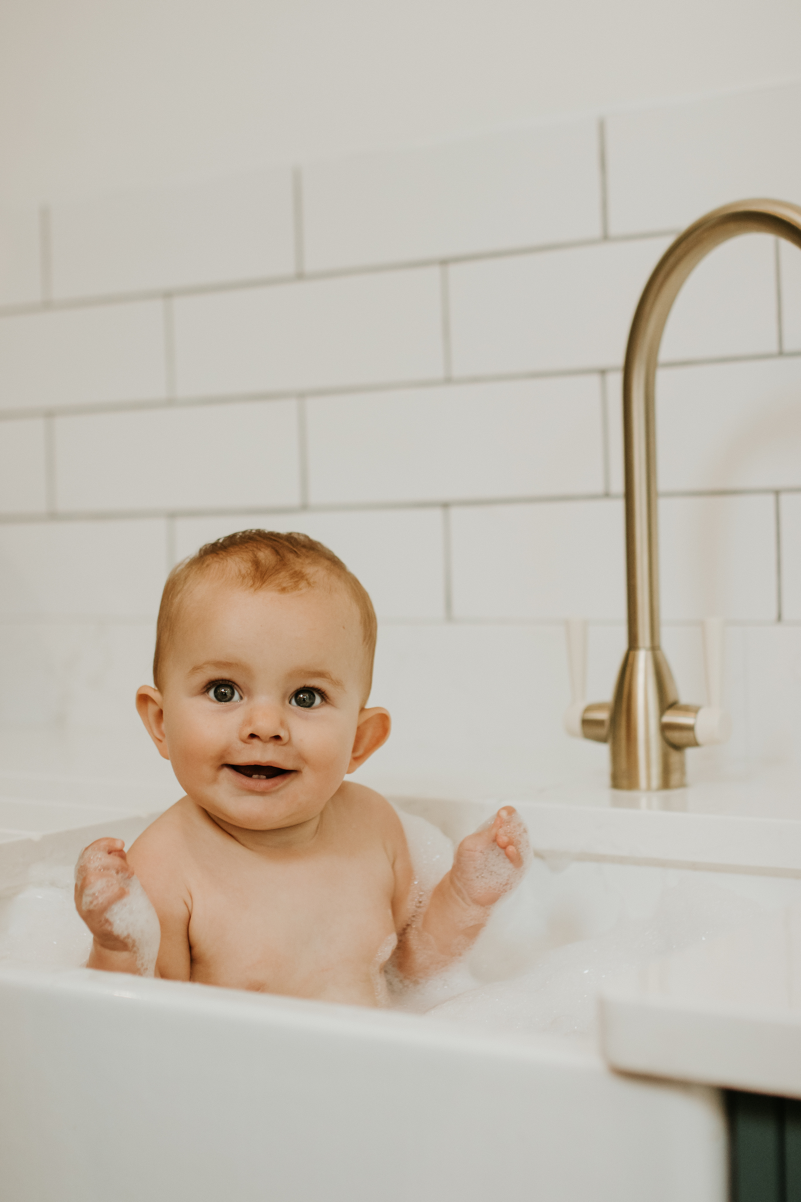 In home baby photography session. Baby is in the sink having a bath and smiling at the camera. 