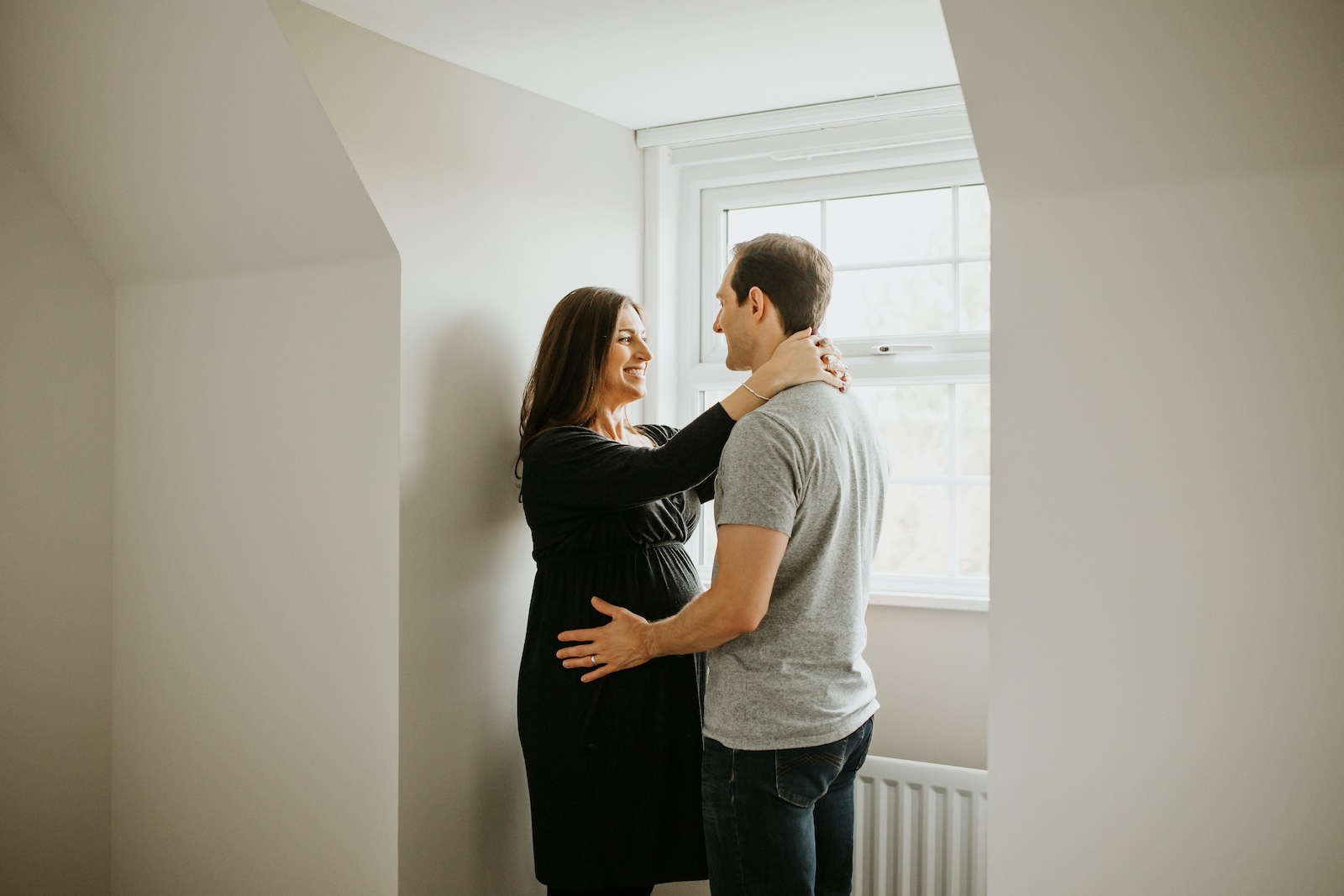 Mum and dad to be standing by bedroom window smiling at each other during pregnancy photoshoot - at home maternity photoshoot