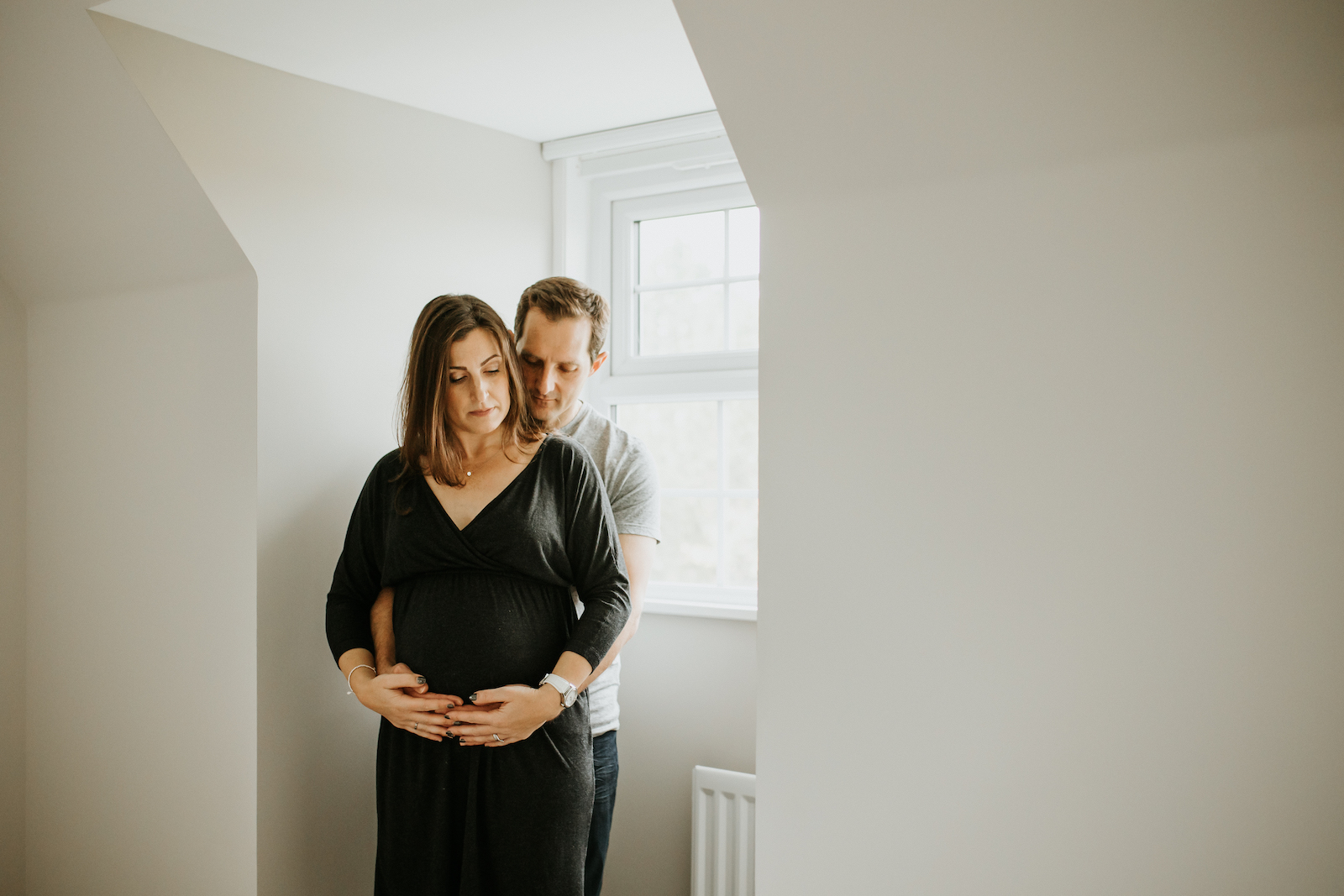 Mum and dad to be standing by bedroom window cradling pregnancy bump - at home maternity photoshoot