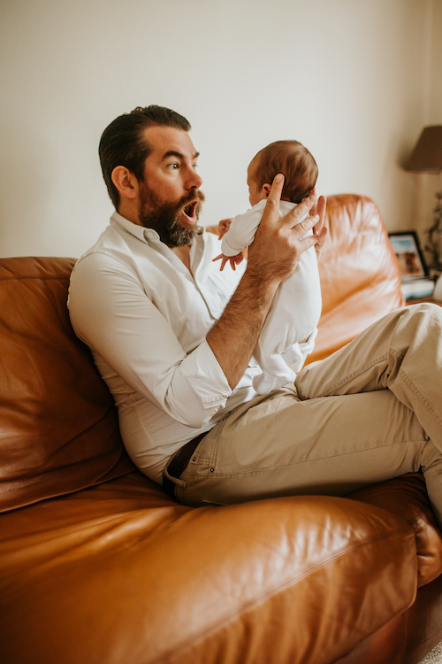 Dad in white shirt and cargo trousers sat on brown sofa holding newborn baby